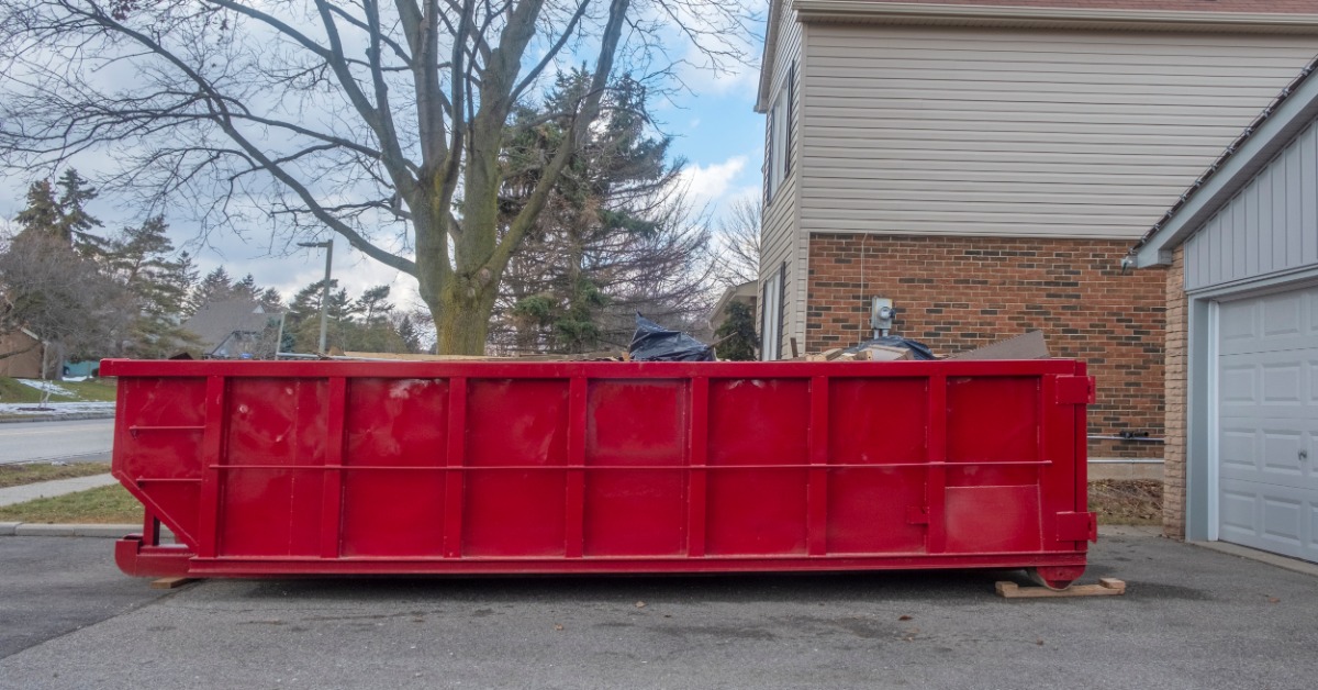 A red dumpster on a driveway next to a home with a white garage door and a large tree in the back on a cloudy day.